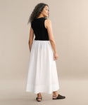 Wearing the Lauren Skirt, a person with long curly hair in a sleeveless black top stands barefoot against a beige background. Effortlessly chic, the Lauren Skirt shines as a true wardrobe staple.