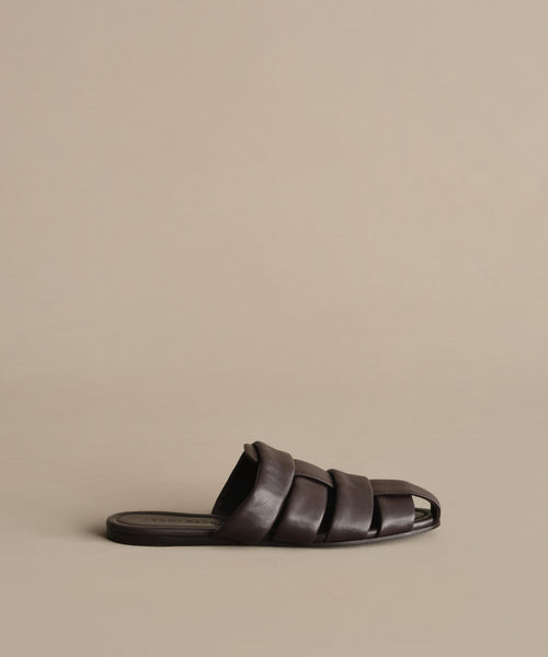 The Leather Camilla Mule is a dark brown, fisherman-inspired slide featuring wide woven leather straps and an open toe, shown against a plain beige background.