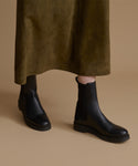 Black leather Chelsea boots worn with an olive green skirt, shown from the knees down against a solid beige background.