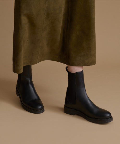 Black leather Chelsea boots worn with an olive green skirt, shown from the knees down against a solid beige background.