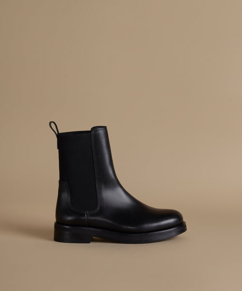 A single black leather Chelsea boot with a black elastic side panel and pull tab, shown in profile against a solid beige background.