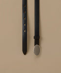 A black leather belt laid flat on a warm beige background, showing the strap with adjustment holes on one side and the buckle on the other. The buckle is a large, polished oval with a silver-toned metal border.