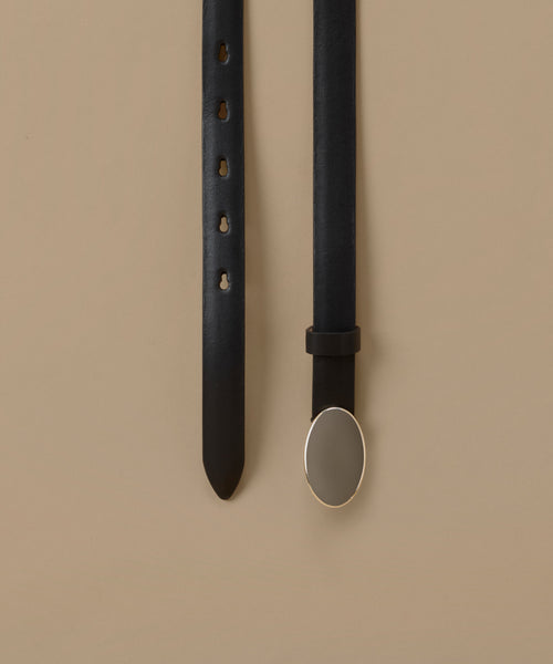 A black leather belt laid flat on a warm beige background, showing the strap with adjustment holes on one side and the buckle on the other. The buckle is a large, polished oval with a silver-toned metal border.
