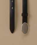 A close-up of a black leather belt shown against a warm beige background, with one vertical strap on the left and another on the right featuring a large oval silver-toned metal buckle.