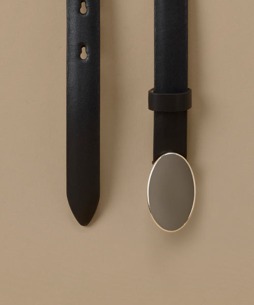 A close-up of a black leather belt shown against a warm beige background, with one vertical strap on the left and another on the right featuring a large oval silver-toned metal buckle.