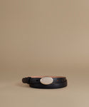 A black leather belt with a large oval white mother-of-pearl style buckle, coiled on a warm beige studio background.