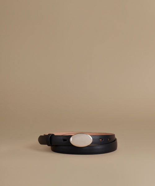 A black leather belt with a large oval white mother-of-pearl style buckle, coiled on a warm beige studio background.