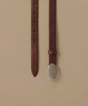A dark brown leather belt with an oval silver-toned metal buckle, shown hanging vertically against a solid beige background.