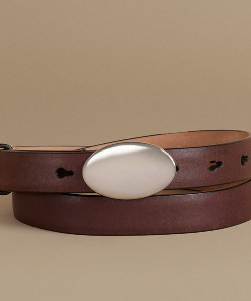 A dark brown leather belt with a large, polished silver oval buckle, neatly coiled on a solid beige background.