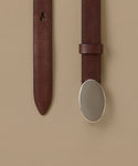 A close-up of a dark brown leather belt with a large, polished silver oval buckle, shown against a solid beige background.