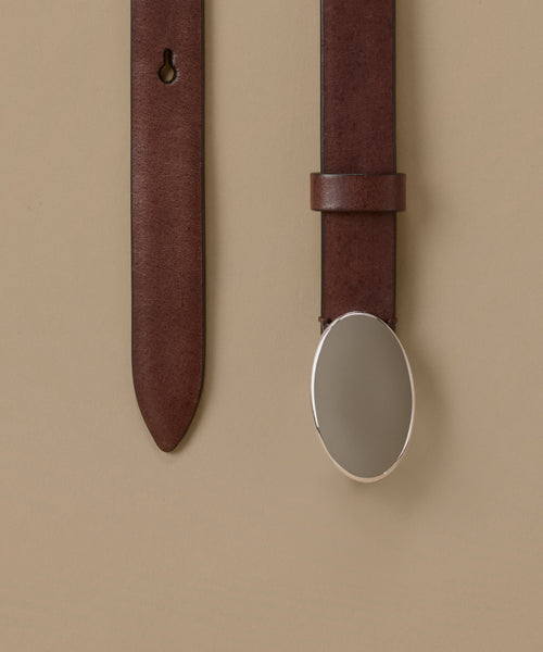 A close-up of a dark brown leather belt with a large, polished silver oval buckle, shown against a solid beige background.