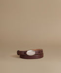 A dark brown leather belt with a large, polished silver oval buckle, neatly coiled on a warm beige studio background.