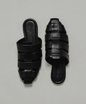 The Leather Camilla Mule is a pair of black leather slip-on sandals with wide interwoven straps and flat soles, displayed on a plain light background.