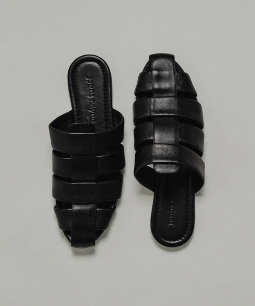 The Leather Camilla Mule is a pair of black leather slip-on sandals with wide interwoven straps and flat soles, displayed on a plain light background.