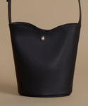 A black pebbled leather bucket bag with a small silver teardrop-shaped metal accent on the front, set against a solid beige background.