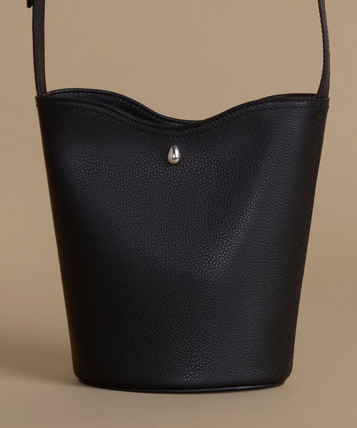A black pebbled leather bucket bag with a small silver teardrop-shaped metal accent on the front, set against a solid beige background.