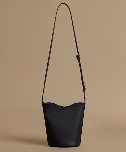 A black leather bucket bag with a long shoulder strap, photographed against a solid beige background.