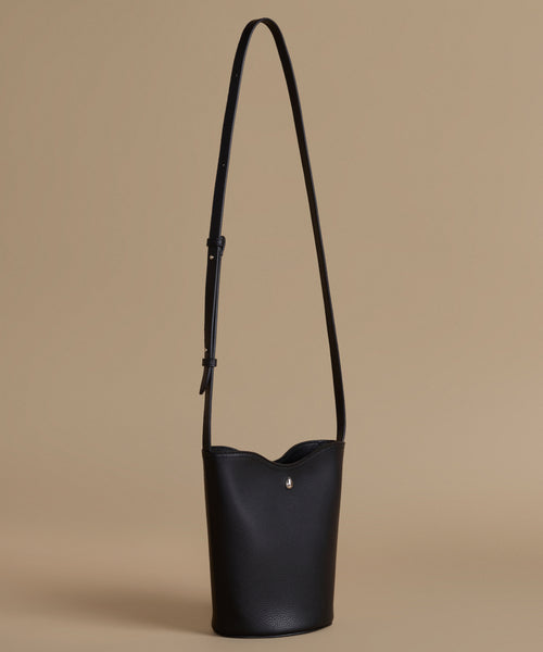 A black leather bucket bag with a long shoulder strap and a small silver logo charm, shown against a solid beige background.