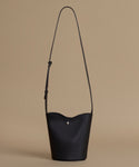 A black leather bucket bag with a long shoulder strap and a small silver clasp, set against a solid beige background.
