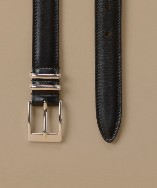 A close-up studio shot of a black leather watch strap laid flat on a warm beige background, showing the stitched strap, silver-toned metal buckle, and adjustment holes.