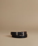 A black leather belt with a silver-tone buckle, neatly coiled on a warm beige studio background.