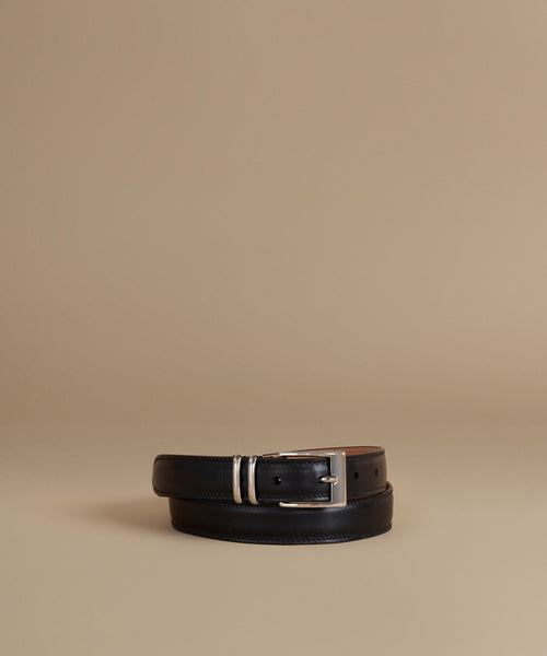 A black leather belt with a silver-tone buckle, neatly coiled on a warm beige studio background.