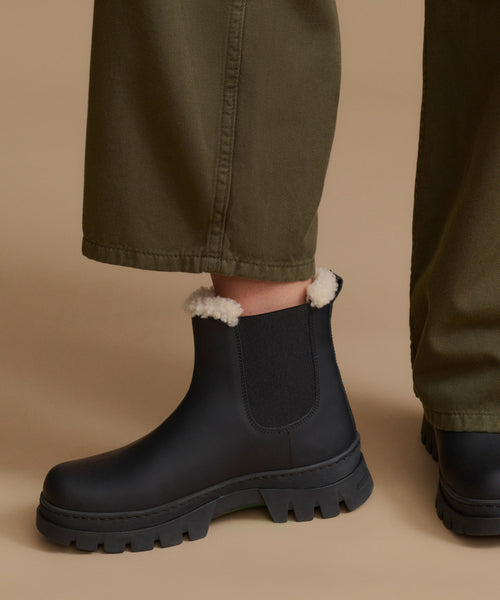 Black Chelsea-style ankle boots with white sherpa lining visible at the collar, worn with olive green trousers against a warm beige studio background.