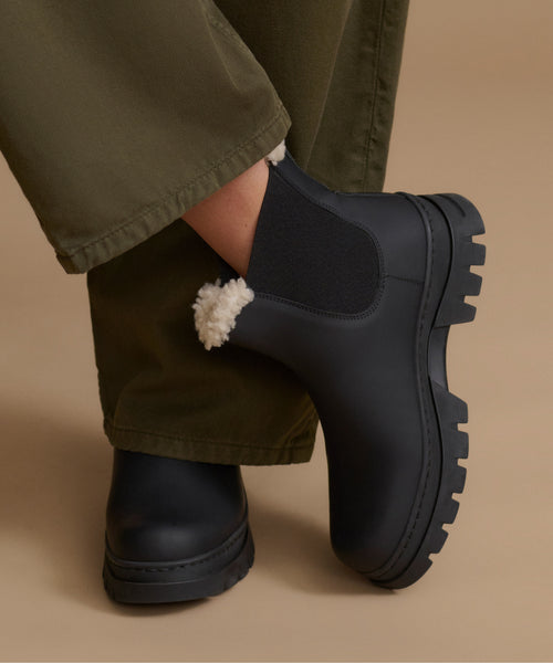 A close-up of a person wearing black Chelsea-style boots with a white sherpa lining, paired with olive green pants, against a solid beige background.