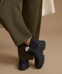 A close-up of a person wearing black Chelsea-style boots with a white faux-fur trim and olive green pants, set against a warm beige background.