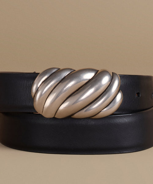 A black leather belt with a large, sculptural metallic silver knot-like buckle, coiled on a warm beige background.