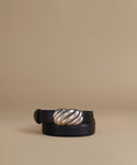 A black leather belt with a large, sculptural twisted silver metal buckle, coiled on a warm beige background.