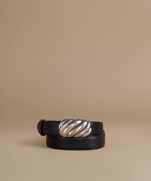 A black leather belt with a large, sculptural twisted silver metal buckle, coiled on a warm beige background.