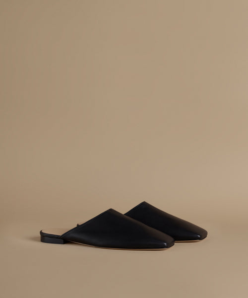 A pair of black leather mule flats with a pointed toe, displayed on a warm beige studio background.