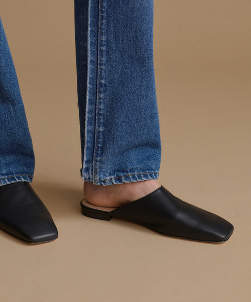 Black leather square-toe mule flats worn with blue jeans, shown from the side against a solid tan background.