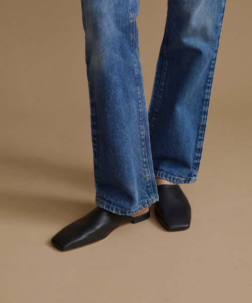 A person wearing blue denim jeans and black square-toe mule flats, standing against a plain warm beige studio background.