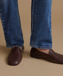 A close-up of a person's legs wearing dark brown leather loafers and blue jeans, standing against a solid beige background.