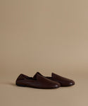 A pair of dark brown leather slip-on loafers with a pointed toe, displayed on a warm beige studio background.