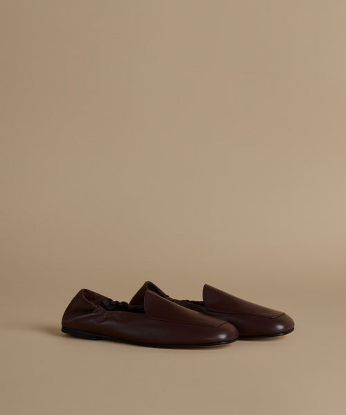 A pair of dark brown leather slip-on loafers with a pointed toe, displayed on a warm beige studio background.