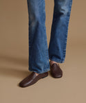 A person wearing blue jeans and dark brown leather loafers, shown from the knees down against a plain warm beige studio background.
