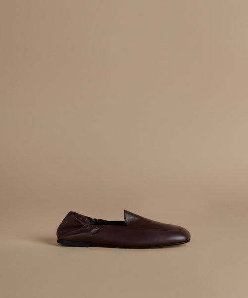 A single dark brown leather slip-on loafer shoe shown in profile against a solid beige studio background.