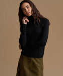 A woman modeling a black long-sleeve mock neck top with a brown skirt, posed against a solid warm beige studio background.
