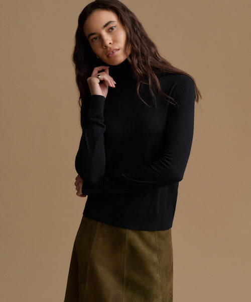 A woman modeling a black long-sleeve mock neck top with a brown skirt, posed against a solid warm beige studio background.