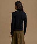 A woman shown from the back wearing a black turtleneck sweater and an olive green skirt, set against a solid warm beige background.