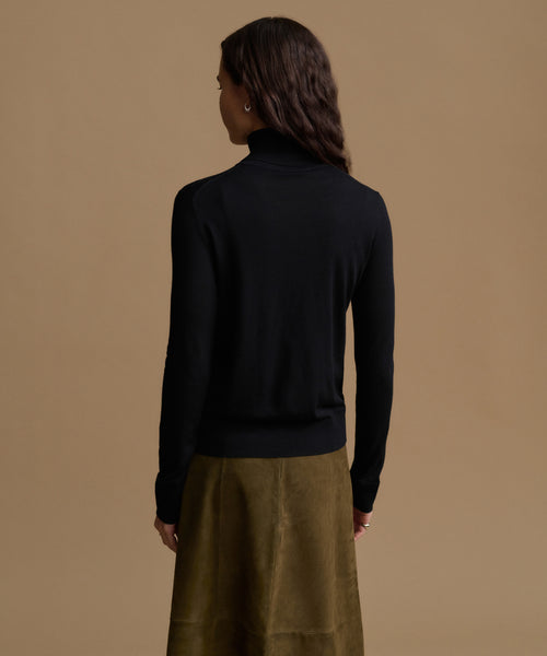 A woman shown from the back wearing a black turtleneck sweater and an olive green skirt, set against a solid warm beige background.