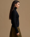 A woman in side profile wearing a black long-sleeve turtleneck sweater and an olive green skirt, set against a solid warm tan studio background.