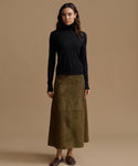 A woman stands against a plain warm beige studio background wearing a black long-sleeve turtleneck sweater and a knee-length olive green skirt with large front patch pockets, paired with dark brown loafers.