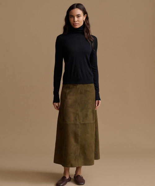 A woman stands against a plain warm beige studio background wearing a black long-sleeve turtleneck sweater and a knee-length olive green skirt with large front patch pockets, paired with dark brown loafers.