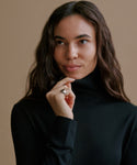 A woman wearing a black turtleneck sweater, posing against a solid beige background.