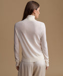 A woman shown from the back wearing a cream-colored knit mock neck sweater and matching knit trousers against a solid beige studio background.