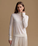 A woman modeling a cream-colored, long-sleeve mock neck knit sweater and matching trousers against a solid beige studio background.